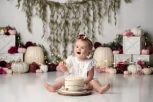 A baby girl is sitting in front of a cake in front of pumpkins.