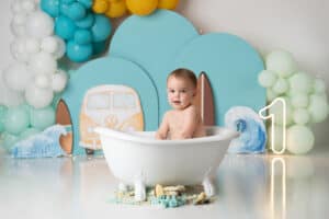 A baby is sitting in a bathtub in front of balloons.
