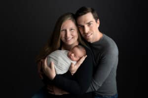 A man and woman holding a newborn baby on a black background.