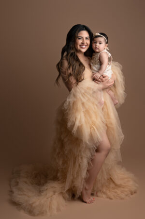 A woman in a tulle dress posing with her baby on a brown background.