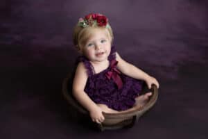 A baby girl sitting in a wooden bowl on a purple background.
