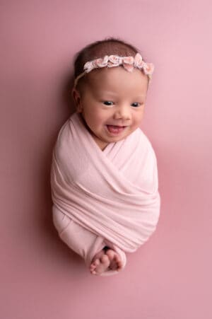 A baby girl wrapped in a pink swaddle on a pink background.