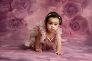 A baby girl in a pink dress crawling on a pink background.