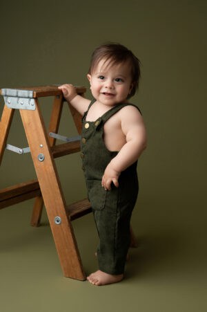 A baby boy in overalls standing on a ladder.
