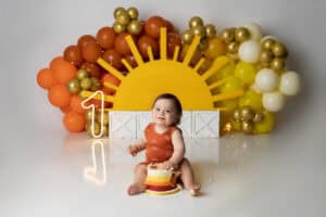 A baby sitting in front of balloons and a sun.