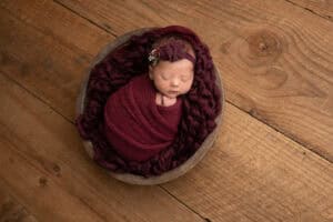 A newborn girl wrapped in a burgundy blanket on a wooden floor.