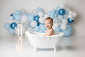 A baby boy sitting in a bathtub with blue balloons.