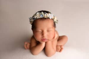 A baby girl wearing a flower crown laying down on a white background.