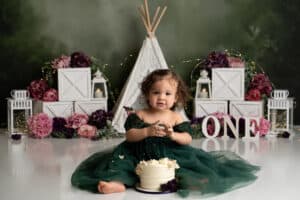 A baby girl in a green dress sits in front of a cake.