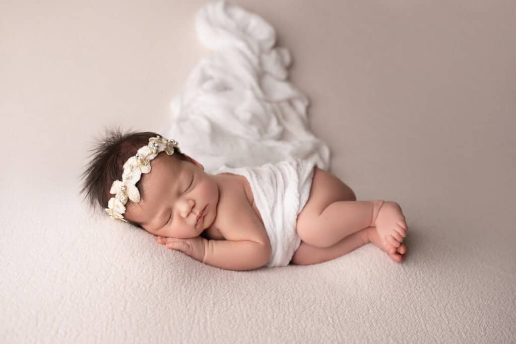 A newborn girl wearing a flower headband laying on a white background.