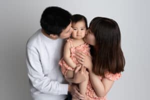 A young asian couple kissing their baby girl in front of a white background.