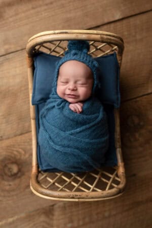 A baby boy in a blue hat is sitting in a wicker basket.