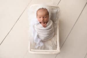 A newborn baby boy in a white blanket laying on a wooden floor.