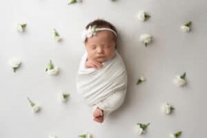 A newborn girl wrapped in a white blanket surrounded by white flowers.