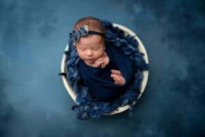 A newborn girl laying in a bowl on a blue background.
