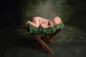 A newborn sleeping on a wooden chair in a green blanket.