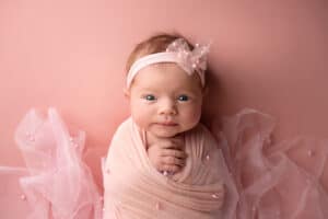 A baby girl in a pink wrap posing on a pink background.