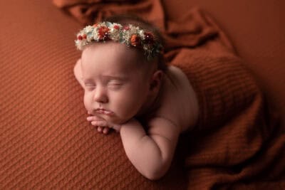 A baby girl wearing a flower crown is laying on a brown blanket.