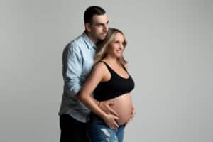 A pregnant couple posing in front of a gray background.