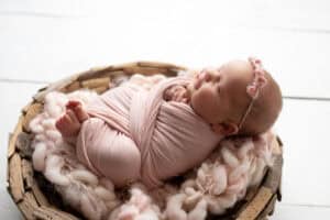 A newborn girl in a pink wrap laying in a basket.
