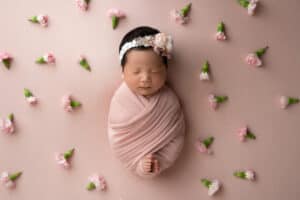 A newborn girl wrapped in a pink blanket surrounded by flowers.