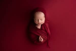 A newborn boy wrapped in a red blanket on a red background.