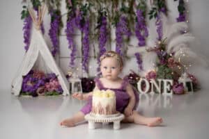 A baby girl in a purple dress sitting in front of a cake.