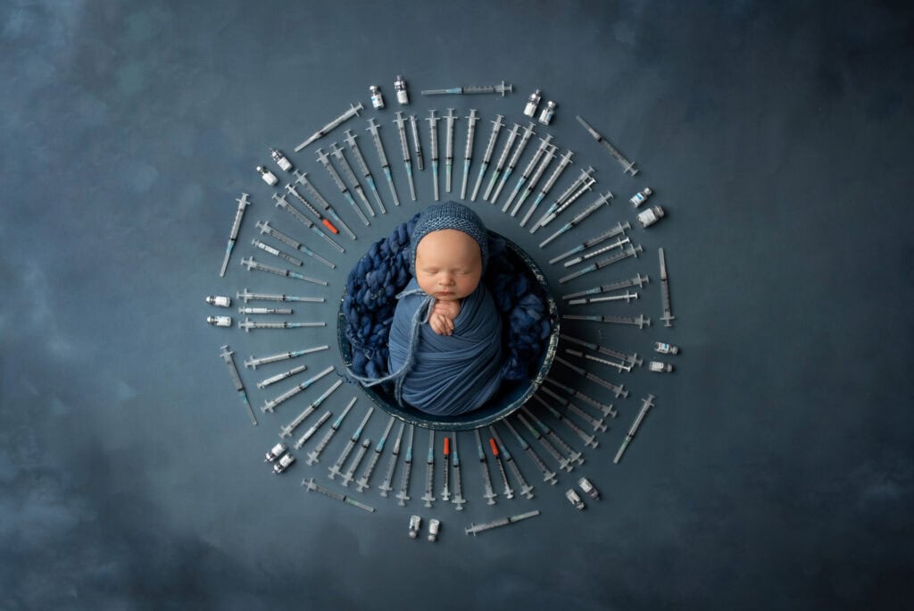 A newborn baby in a blue blanket surrounded by a circle of syringes.