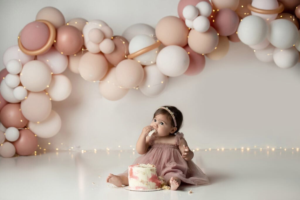 A baby girl sitting in front of a pink and white balloon arch.