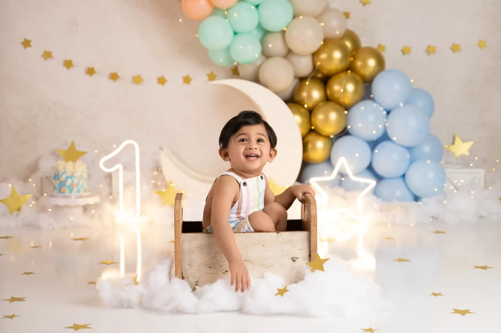 a joyful baby sits in a decorative wooden box surrounded by balloons, sparkly stars, and a "1" light sign, celebrating their first birthday in a festive setup.
