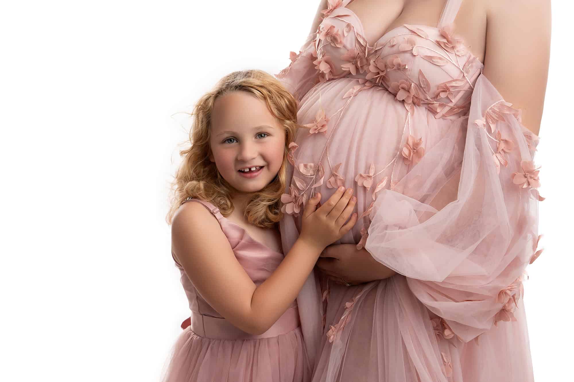 a young girl in a pink dress smiles while embracing a pregnant woman's belly, which is clad in a flowing pink gown with floral details, celebrating hot mom summer. the background is white.