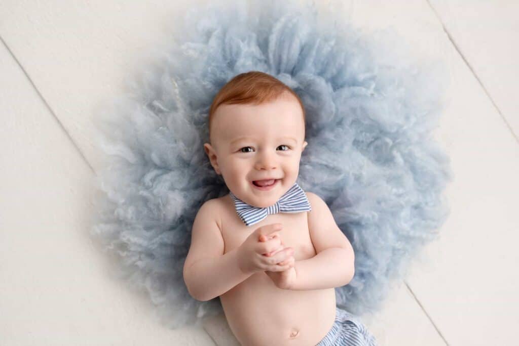 a joyful baby with a light blue bow tie lies on a fluffy, blue tulle fabric during an infant photoshoot, smiling and clasping hands.