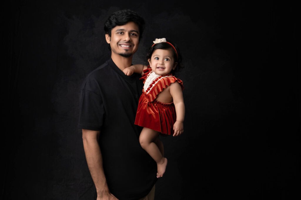 a man wearing a black shirt is seen holding a smiling baby dressed in a red dress with a white bow on her head. both are posing against a dark background, capturing the perfect moment of baby photography.