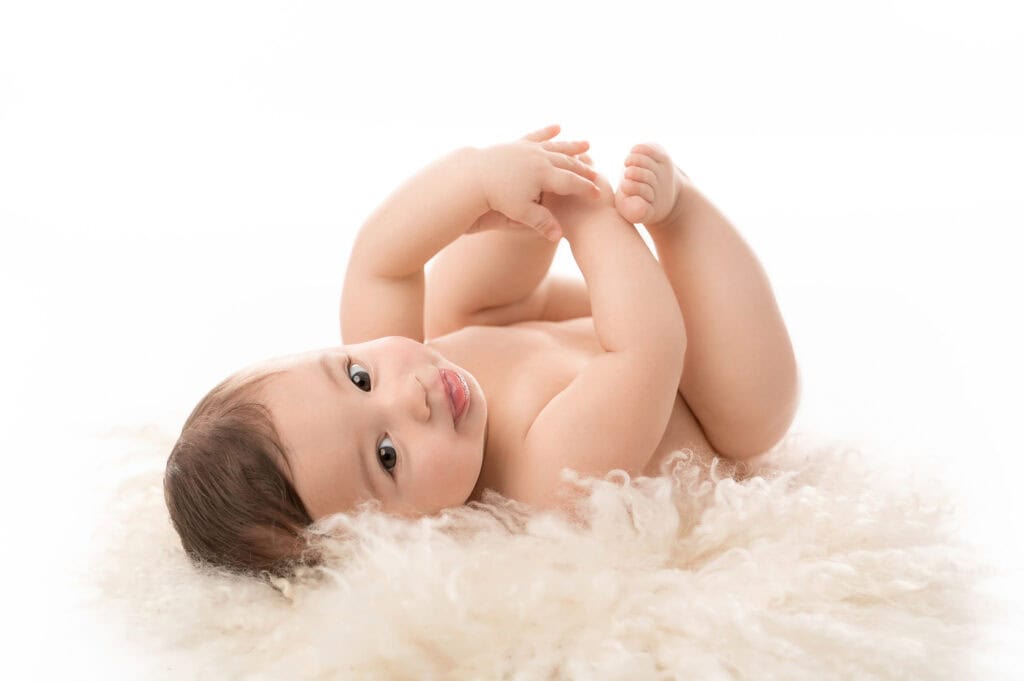 a baby lies on a fluffy white blanket, holding its feet with both hands. the baby looks directly at the camera with wide eyes and a slight smile. the background is a soft, light color, enhancing the gentle, serene atmosphere of this newborn pictures session.