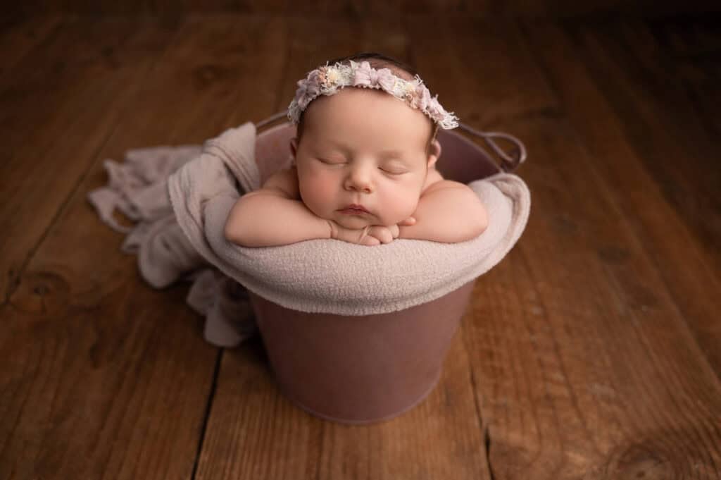 a baby peacefully sleeps with their head resting on folded arms inside a pink bucket lined with a soft beige blanket. they wear a delicate floral headband. the background is a rustic wooden floor, captured beautifully by a sacramento maternity and newborn photographer at their renowned studio.
