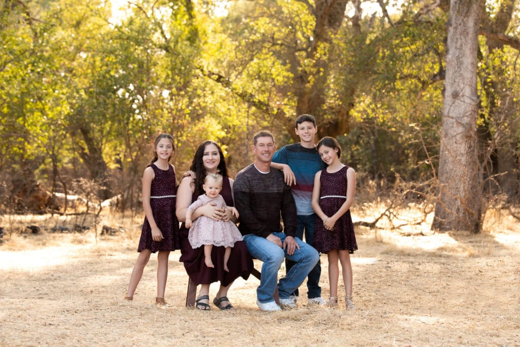 a family of six poses outdoors in a sunlit, wooded area. the group includes a man, a woman, an infant, and three young girls. the woman is holding the infant, and everyone is wearing shades of burgundy and brown, smiling warmly at the camera a perfect moment of family & child photography.
