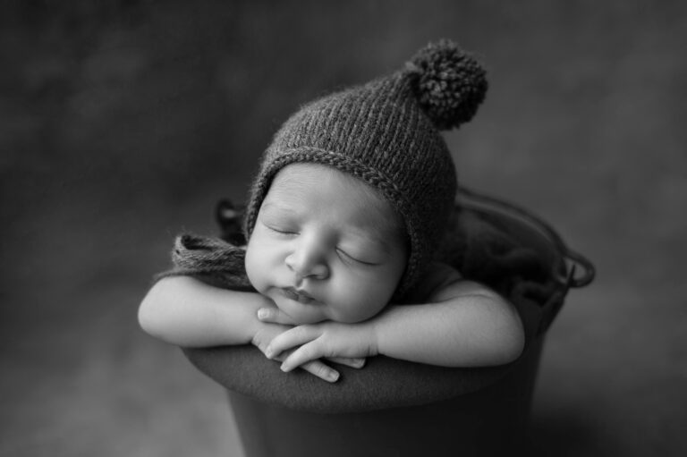 black and white photo of a sleeping baby wearing a knit hat with a pom pom, resting their head and arms on the edge of a round bucket. the background is softly blurred, drawing attention to the baby's peaceful expression, captured perfectly by our sacramento baby portrait studio.