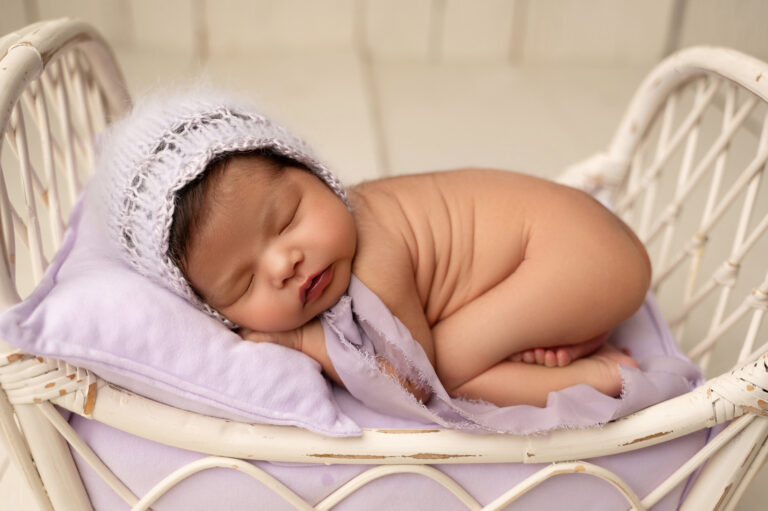 a newborn baby peacefully sleeps on a delicate white wicker bed with a soft lavender cushion and blanket, wearing a cozy white knit cap. the baby is curled up on their side, creating an adorable and serene scene captured beautifully during a sacramento baby photography session.