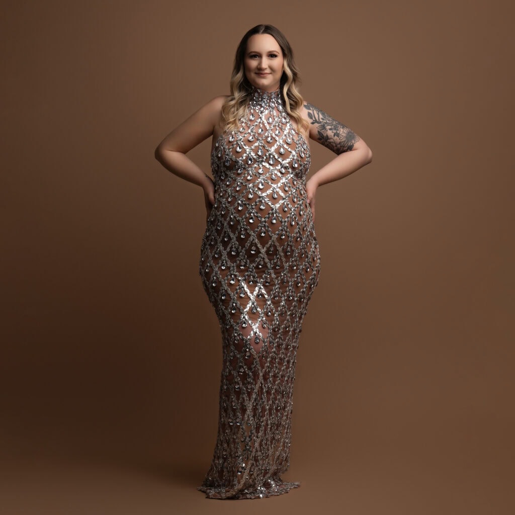 a pregnant woman in a sleeveless, form fitting, sequined gown poses with hands on hips against a plain brown background. she has long, wavy hair and a tattoo on her right arm, embodying the elegance of maternity photography.