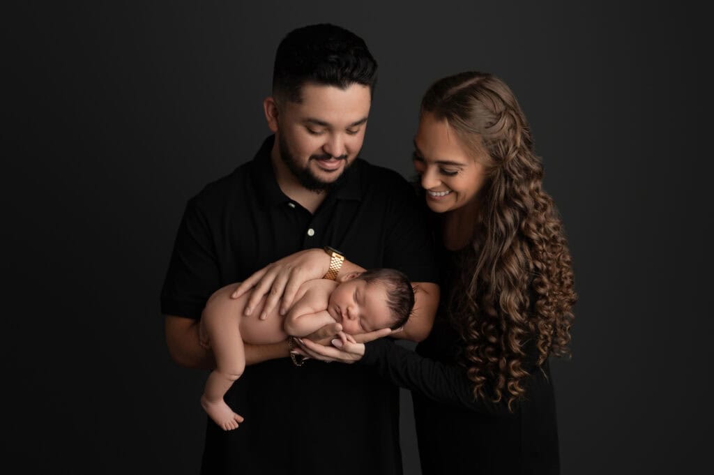 a couple, dressed in black, lovingly cradles a sleeping newborn baby. the man gently holds the baby while the woman, with long curly hair, looks adoringly at the infant. the plain dark background at our sacramento baby portrait studio highlights this tender family moment.