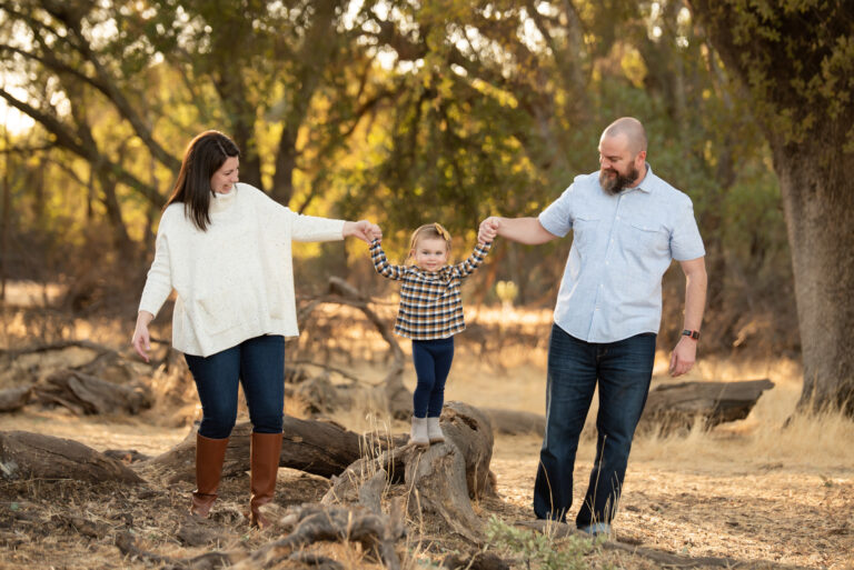 a family of three enjoys an outdoor walk. a woman on the left and a man on the right hold the hands of a young child standing on a fallen tree. the picturesque scene, perfect for family photography, captures them surrounded by a wooded area with autumn foliage.