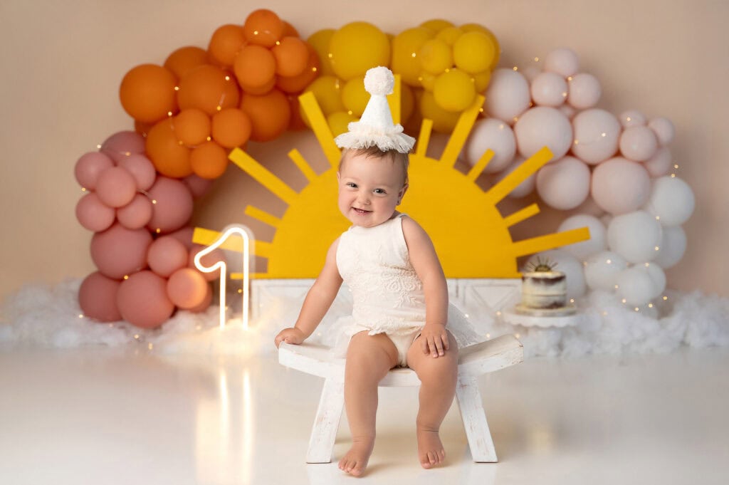 a toddler wearing a white outfit and a party hat sits on a small white bench. the backdrop features an arrangement of balloons in shades of orange, yellow, and white, forming a sunrise design. perfect for cake smash photography, the decorations include a lit number "1" and a small cake ready for the smash.