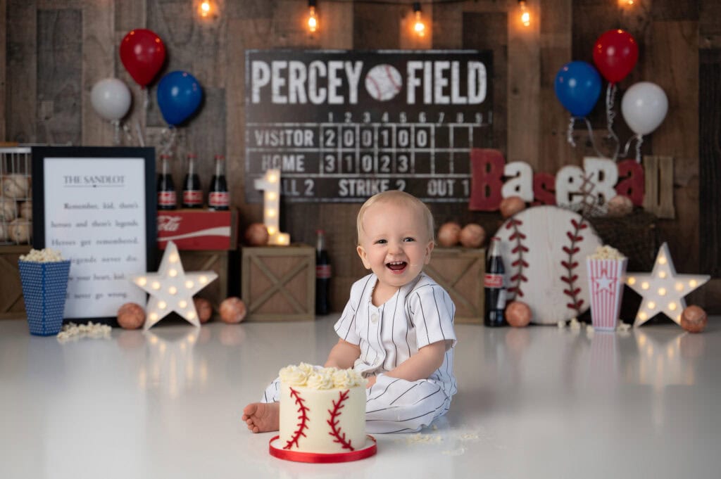 a baby in a baseball themed setting is smiling and sitting in front of a cake decorated like a baseball. the background features lights, balloons, baseballs, a scoreboard, and lettering that spells "baseball." decorative stars and a popcorn box are also visible—a perfect celebration for the new member of the first year club.