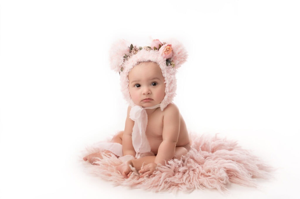 a baby sits on a fluffy pink rug, wearing a soft pink bear themed hat adorned with flowers and a white ribbon around the neck. the background is white and uncluttered, perfectly capturing the essence of newborn photography and highlighting the baby's curious expression.