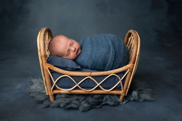 a newborn baby, a proud member of the first year club, is peacefully sleeping in a small, rustic, woven basket crib. the baby is wrapped in a cozy blue blanket with a matching blue pillow supporting its head. the background is a dark, soft, textured fabric, enhancing the tranquil and serene scene.