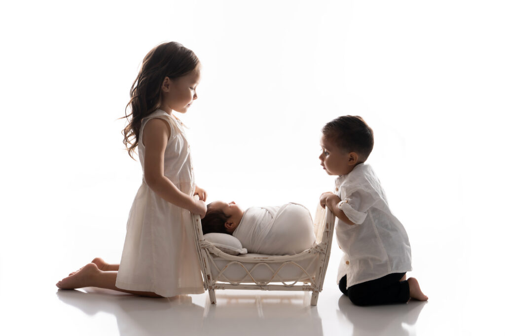 a young girl and boy kneel on the floor, gazing at a baby wrapped in a white blanket, sleeping in a small cradle. the scene is bright and minimalistic, capturing a serene moment reminiscent of an image from 2025.