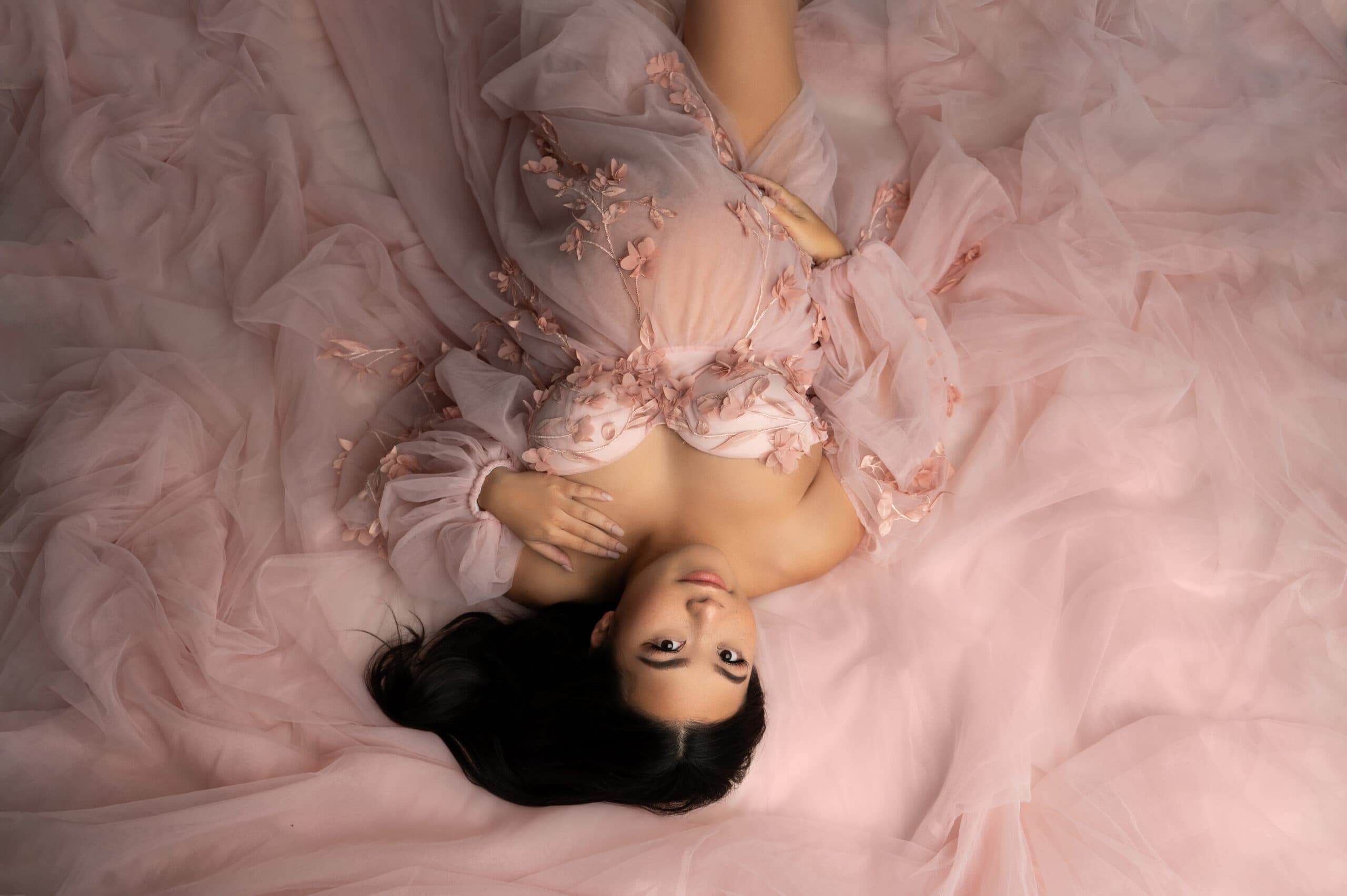 a pregnant woman lies on a soft, pink fabric, embodying the promise of 2025. she wears an elegant, flowing pink dress with floral details, her long dark hair spread out. her hand rests gently on her belly as she gazes upward with a serene expression, much like one savors reading an investment guide.