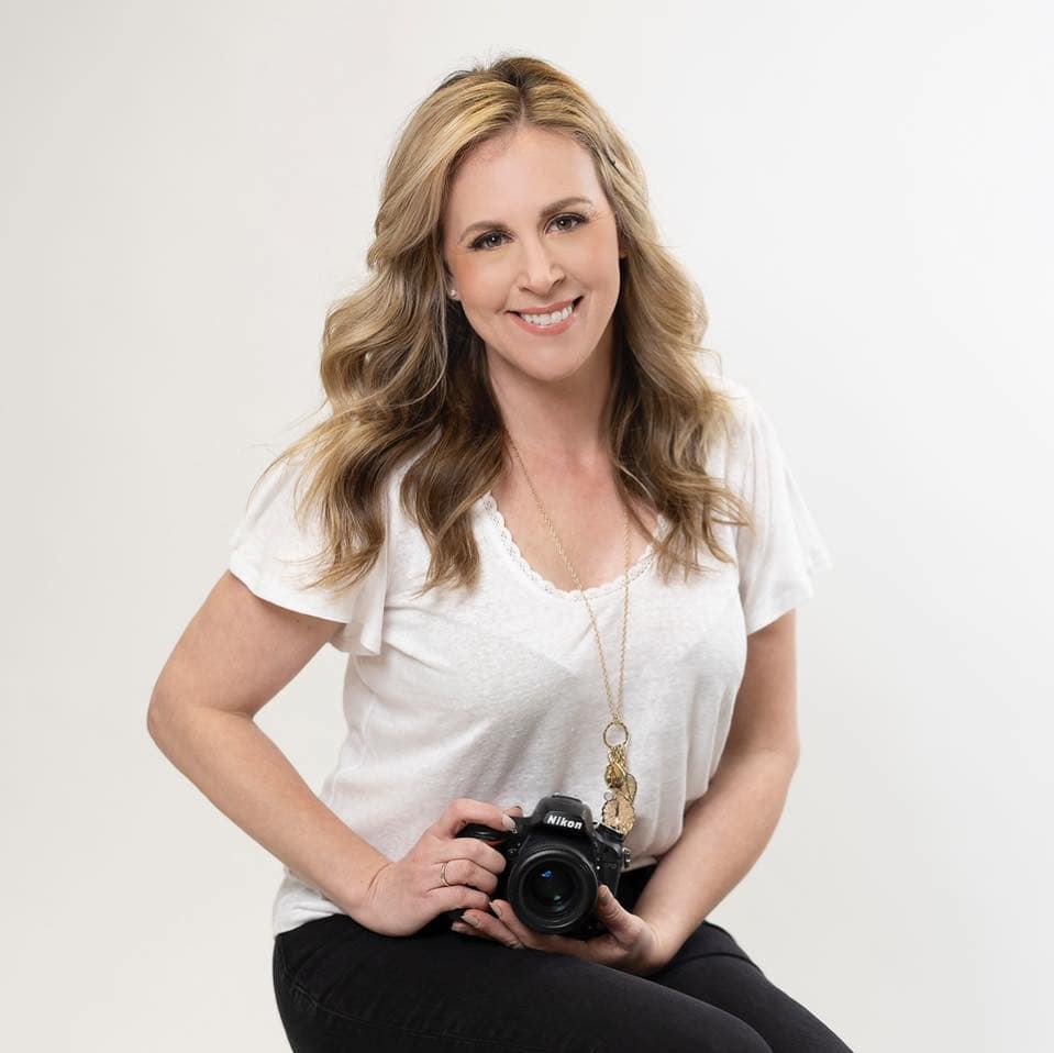 person with long, wavy hair smiles at the camera, holding a nikon camera. they are wearing a white top, black pants, and a necklace with a pendant. the background is plain and white.