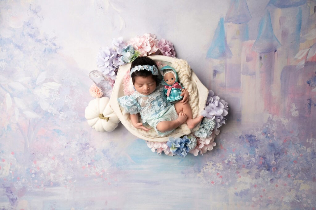 a sleeping baby dressed in blue lace lies in a white basket surrounded by pastel flowers. beside the baby is a small doll, all beautifully captured by a sacramento baby photography expert. the backdrop features a dreamy, painted castle scene in soft pastel colors.