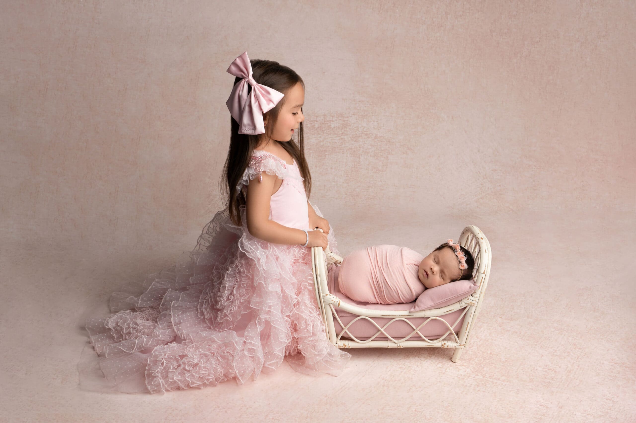 Girl in pink dress beside sleeping baby on bed.
